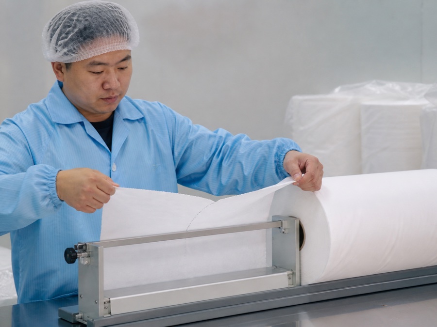 exam couch roll manufacturer — Chinese adult quality inspector in hairnet tearing a white exam couch roll sheet along the perforation using a repeatable