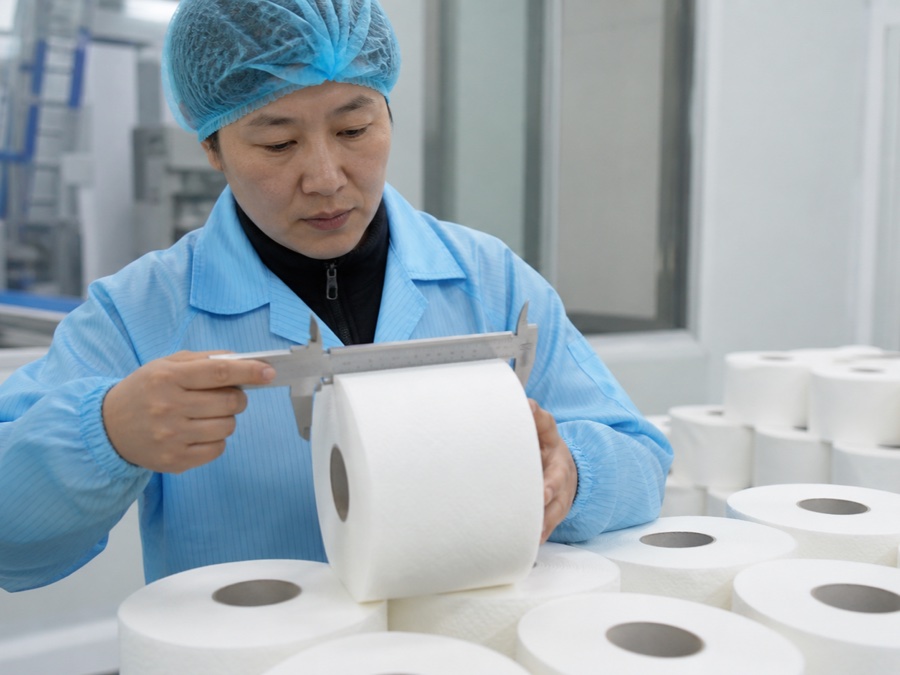 toilet paper supplier China — Chinese adult quality inspector in hairnet measuring toilet roll diameter at a clean packing